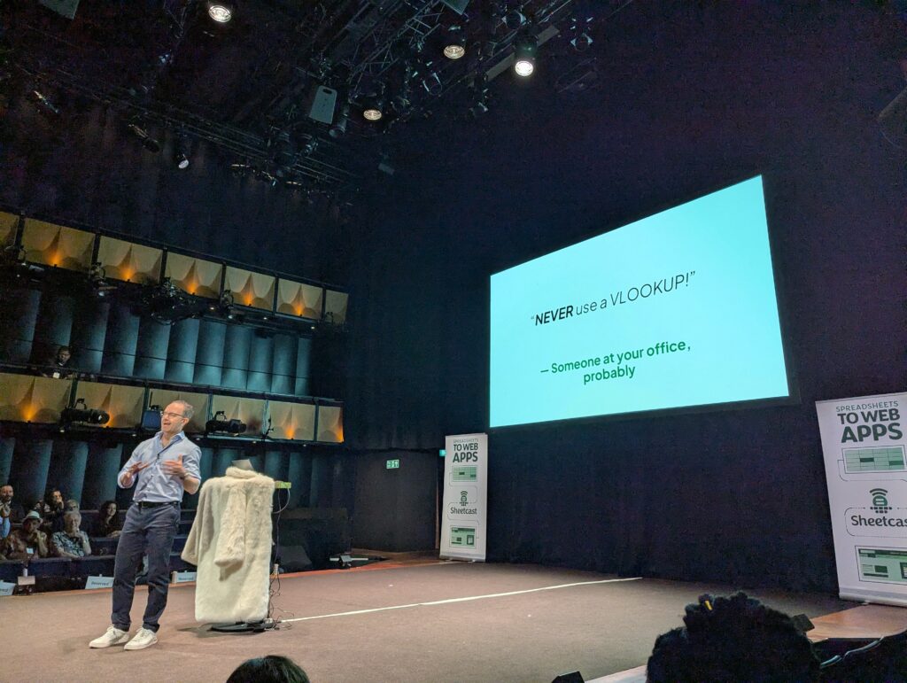 A man is presenting; behind him is a slide that says: "Never use a VLOOKUP!
- someone in your office, probably"
